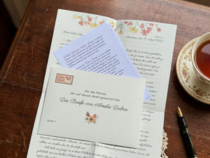 Geöffneter Dein-Brief-Umschlag "Die Briefe von Amélie Dubois" mit Willkommenskarte und Brief auf einem Holztisch neben einer Tasse Tee